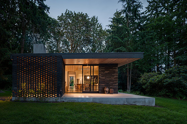Modern single-story home with illuminated interior at dusk, featuring a unique brickwork design and nestled within a tranquil, wooded landscape.